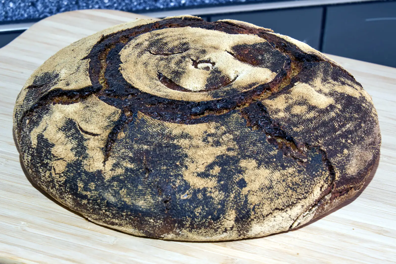 A round loaf of dark sourdough bread dusted with flour on a wooden board.