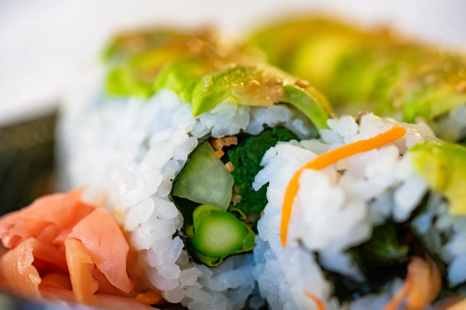 A close-up of a vegetable sushi roll topped with avocado, with pickled ginger.